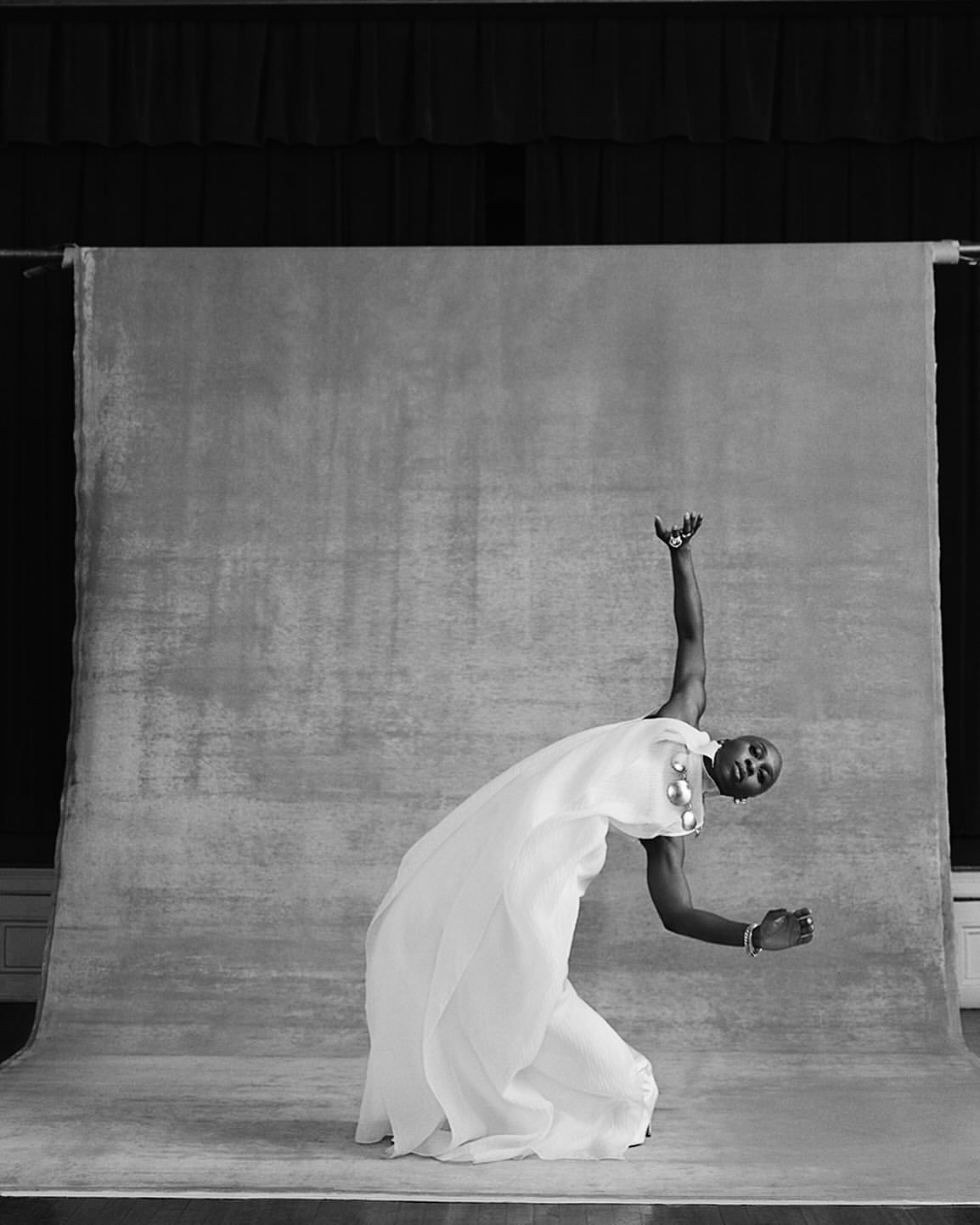 Black and white studio portrait of a person with short hair wearing a flowing white gown, striking a pose with arms extended.