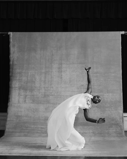 Black and white studio portrait of a person with short hair wearing a flowing white gown, striking a pose with arms extended.
