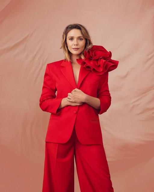 Fashion portrait of Elizabeth Olsen in red suit with ruffled shoulder detail against blush background, shot at eye-level with studio lighting.