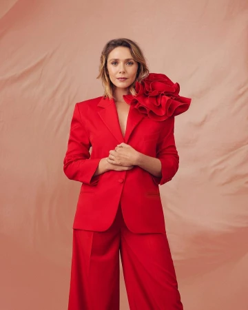 Fashion portrait of Elizabeth Olsen in red suit with ruffled shoulder detail against blush background, shot at eye-level with studio lighting.