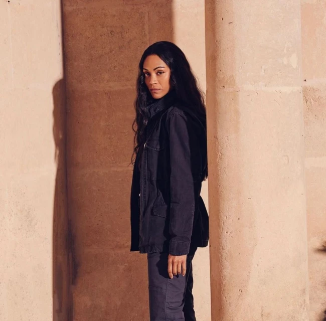 Side profile of an adult female with long dark hair standing against an architectural wall, wearing a dark jacket and pants.