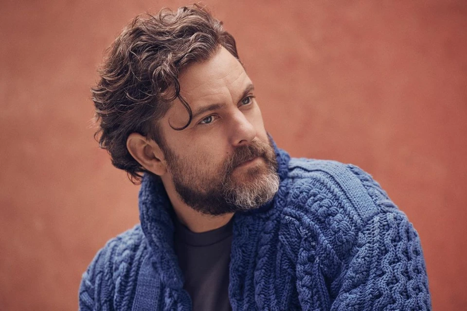 Portrait of an adult male with dark curly hair and a beard, wearing a blue knitted cardigan, set against a warm peach backdrop.