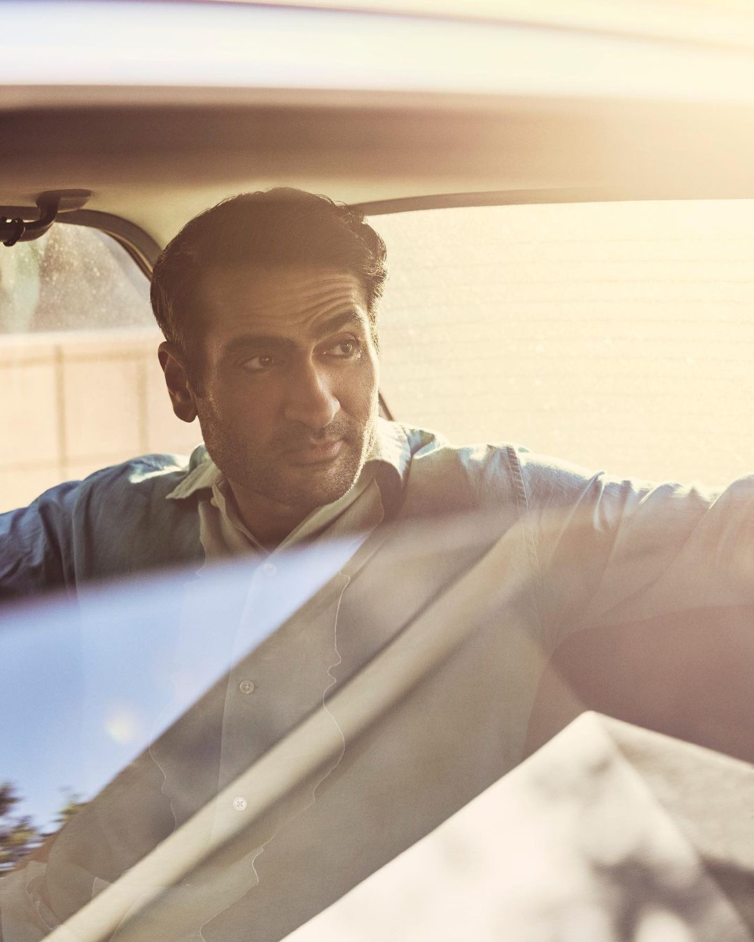 Close-up of a man in a car, wearing a light blue shirt, looking off to the side with a serious expression, bathed in warm sunlight.