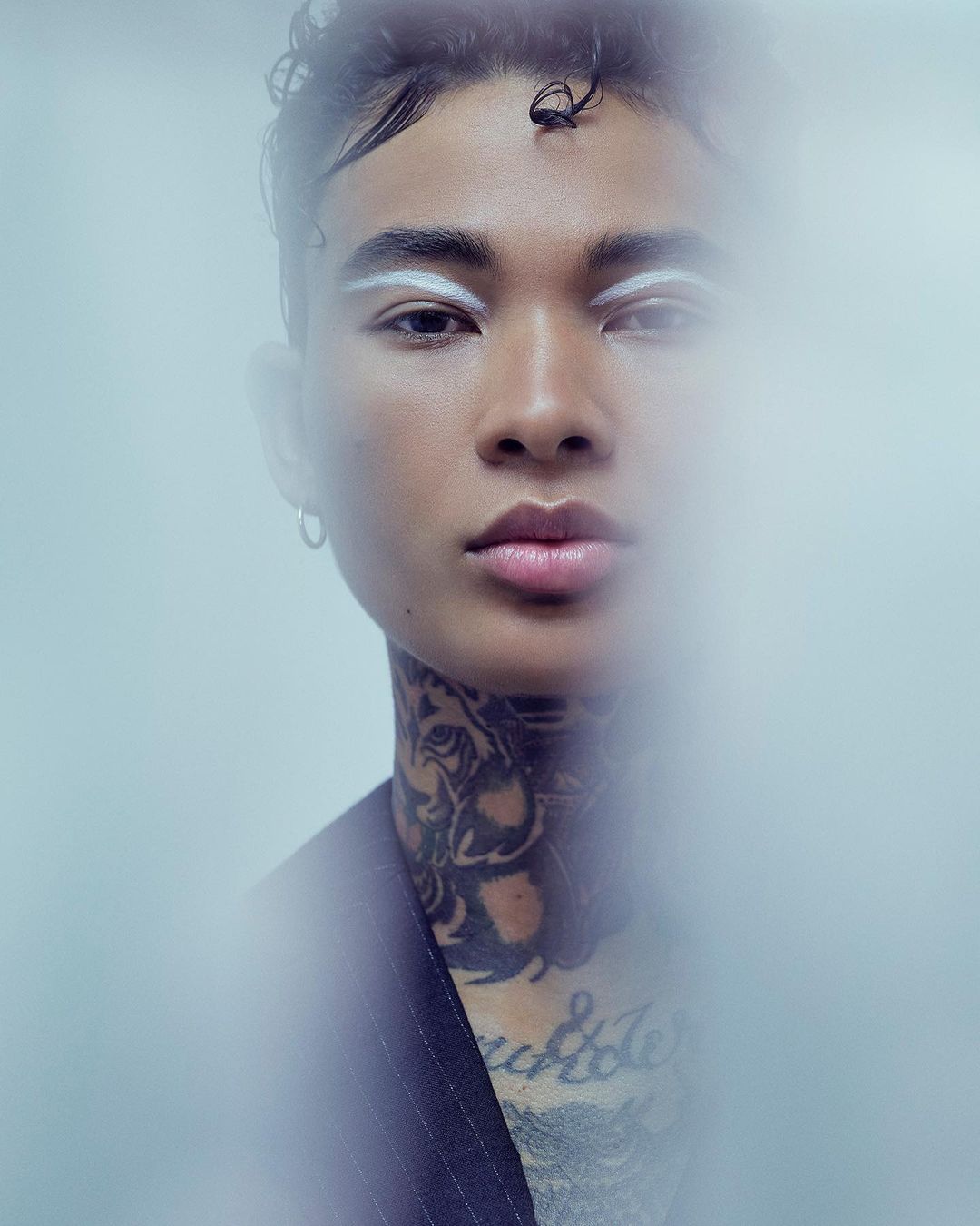 Close-up studio shot of a person with an artistic white eyebrow makeup design, face tattoos, and pinstripe jacket against light background.