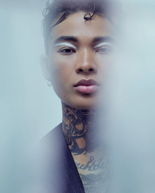 Close-up studio shot of a person with an artistic white eyebrow makeup design, face tattoos, and pinstripe jacket against light background.