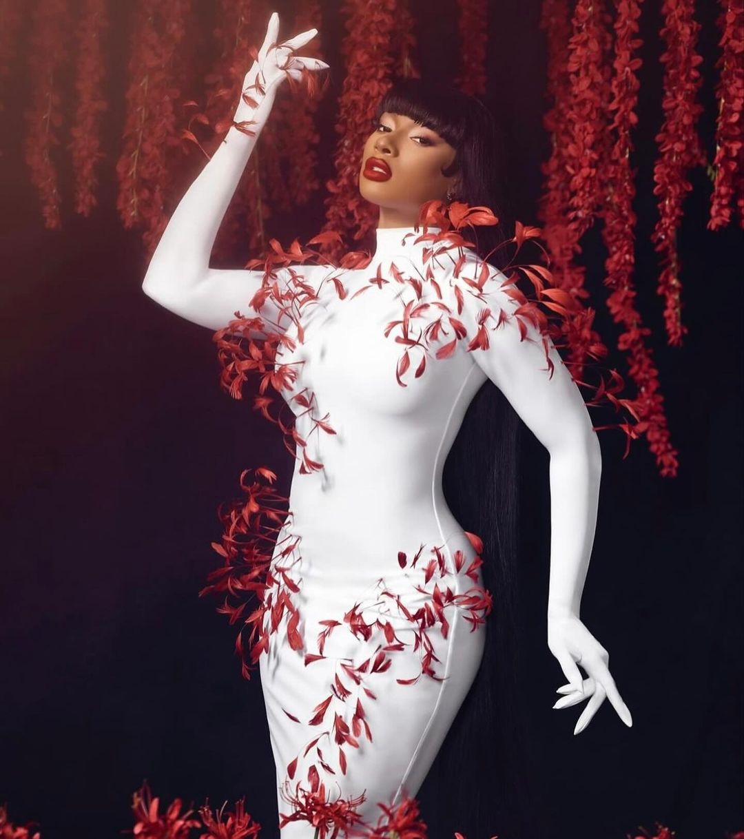 Woman poses in long white dress with red leaf details against a black background with soft lighting, exuding fashion and style.