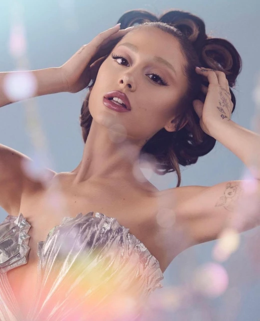 Close-up portrait of Ariana Grande in a silver bustier with hand in hair, against a pastel blue backdrop with bokeh and soft lighting.