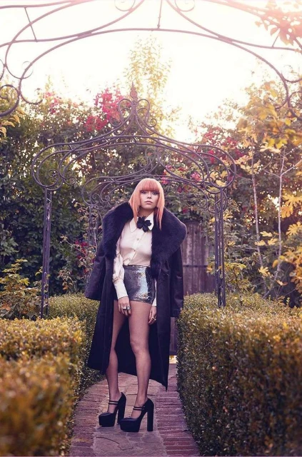 A stylish woman with pink hair wearing a black coat and platform heels stands in an outdoor garden setting with lush greenery.