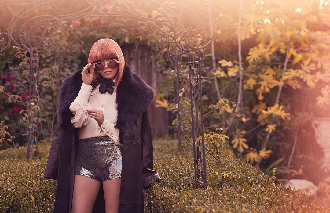 A person with reddish hair wearing large sunglasses, a white blouse, and sparkly shorts stands in front of greenery and a metal archway.