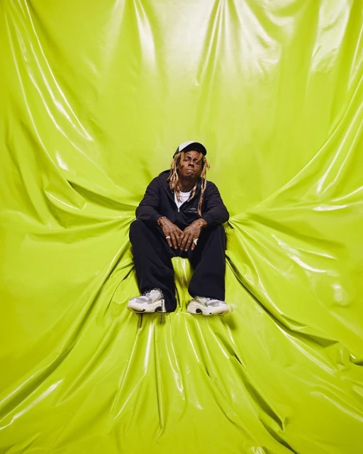 Lil Wayne sitting in front of a bright yellow draped background in a studio. He is wearing a black jacket, pants, cap and white sneakers.