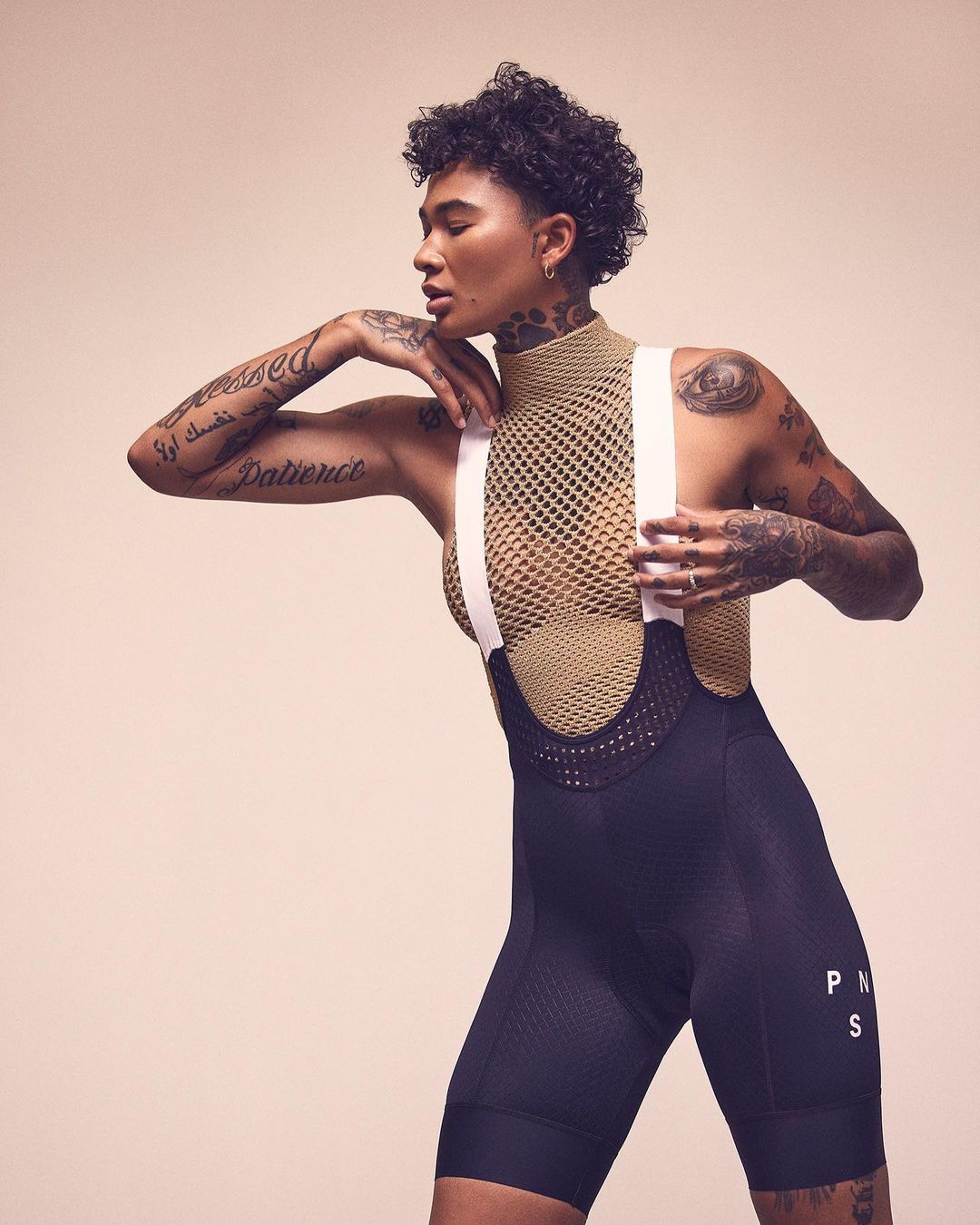 Woman in fitness apparel poses in a studio against a neutral background, showcasing visible tattoos and athletic wear.