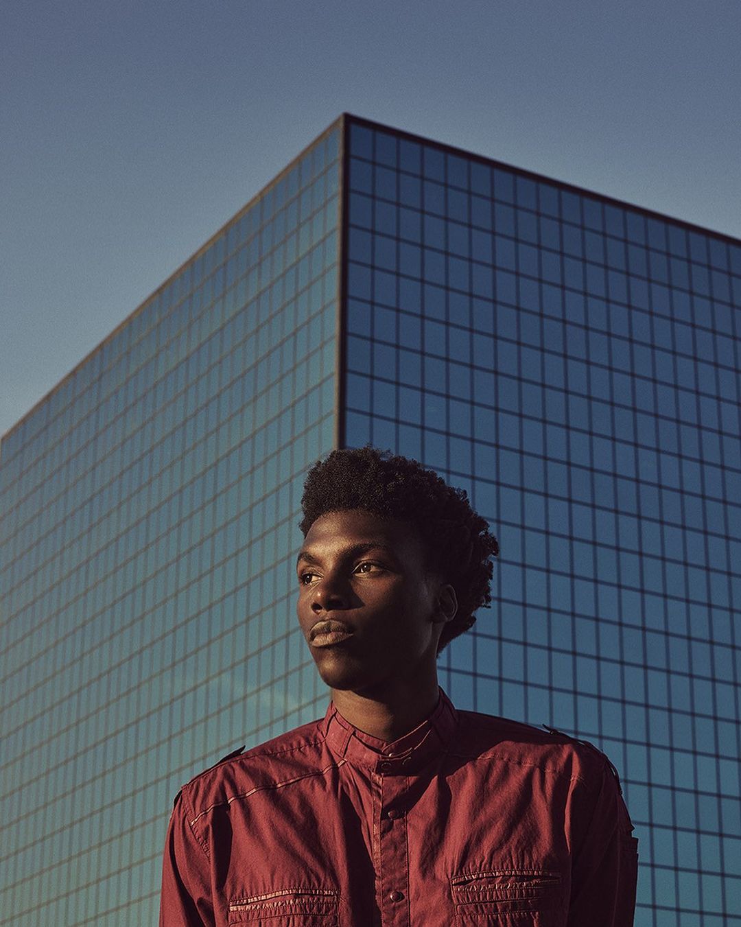 Portrait of an African-American man in a maroon shirt, captured at eye-level with a modern glass building and clear sky in the backdrop.
