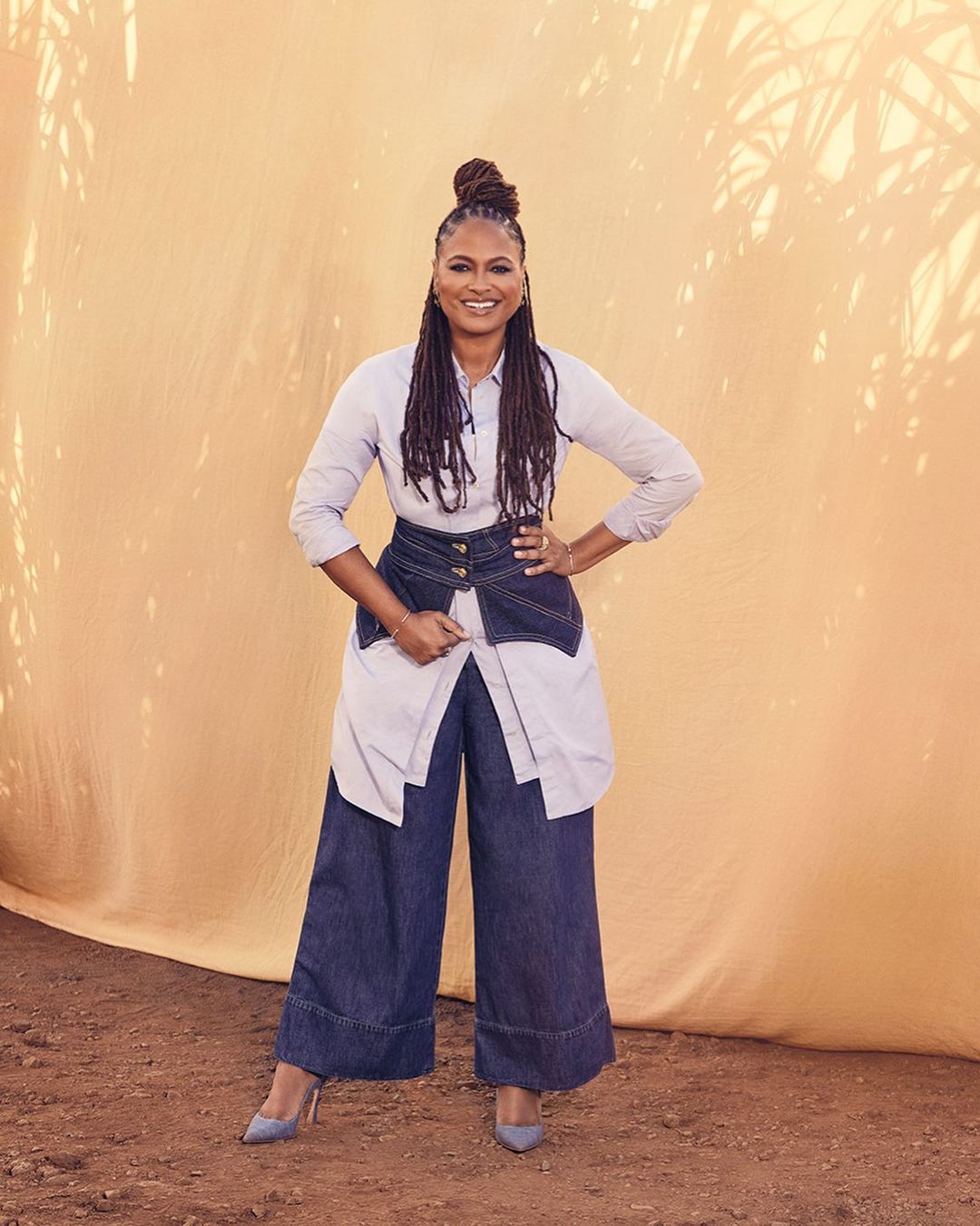 Ava DuVernay in light blue shirt and dark denim wide-leg pants posing against draped yellow background