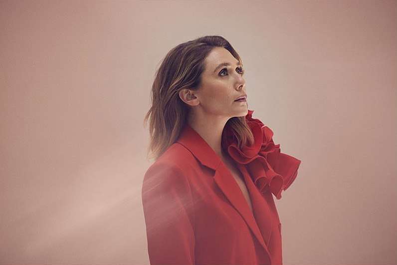 Portrait of a woman wearing a red blazer with a large ruffled shoulder detail, looking up against a peach-colored background.