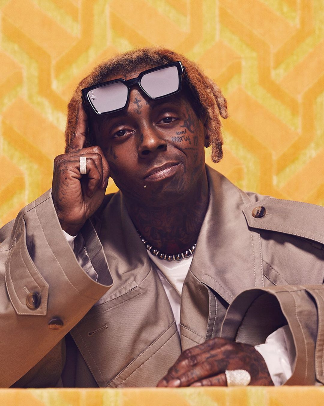 Portrait of Lil Wayne wearing black sunglasses and a tan trench coat against a geometric patterned background.