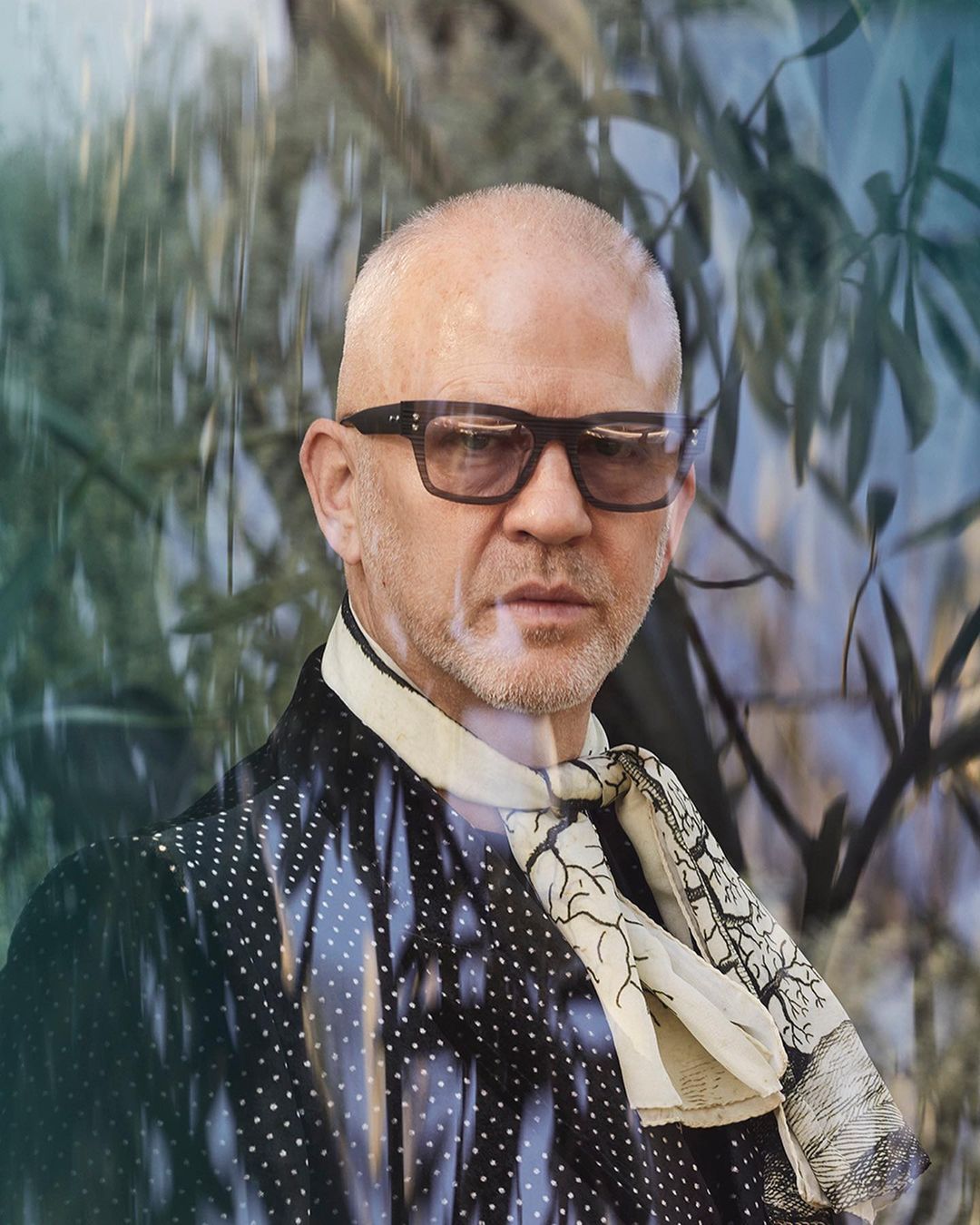 Close-up portrait of a bald man wearing sunglasses, a patterned jacket, and a scarf with a tree design, set against a leafy background.
