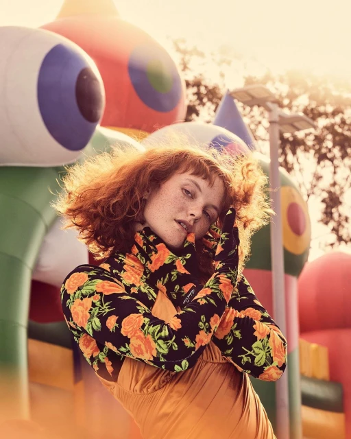 Portrait of a redhead woman in a floral turtleneck and orange dress, posing outdoors with a blurred bouncy castle background in sunlight.