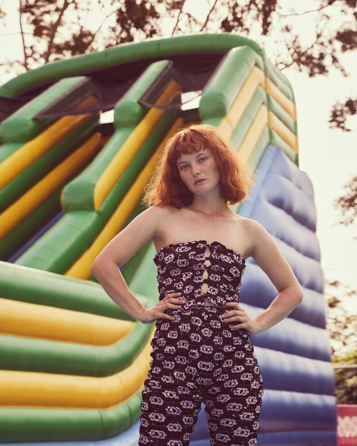 Adult woman with red hair poses in front of inflatable slide wearing strapless patterned top and pants outdoors in soft, diffused lighting.
