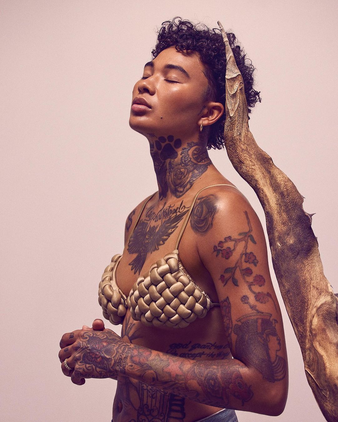 Woman with short curly hair and tattoos poses with a woven bra and a piece of driftwood in a studio with a light pink background.