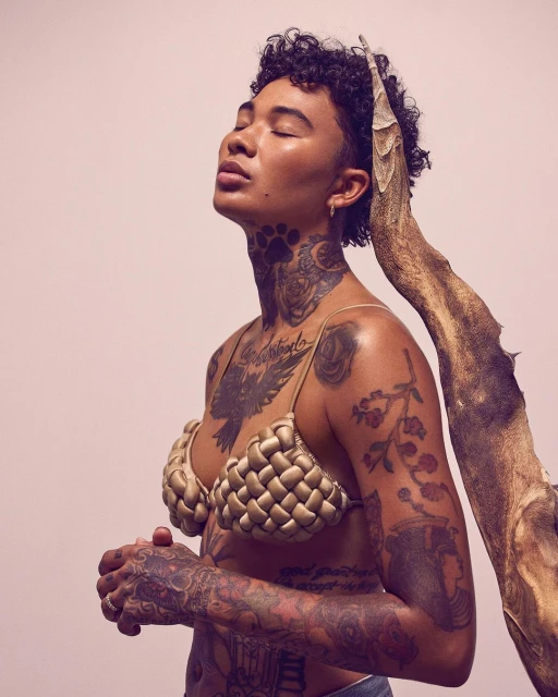 Woman with short curly hair and tattoos poses with a woven bra and a piece of driftwood in a studio with a light pink background.
