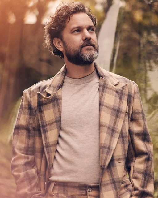 Portrait of a bearded man in a plaid blazer outdoors, looking up with a neutral expression, against a blurred foliage background.