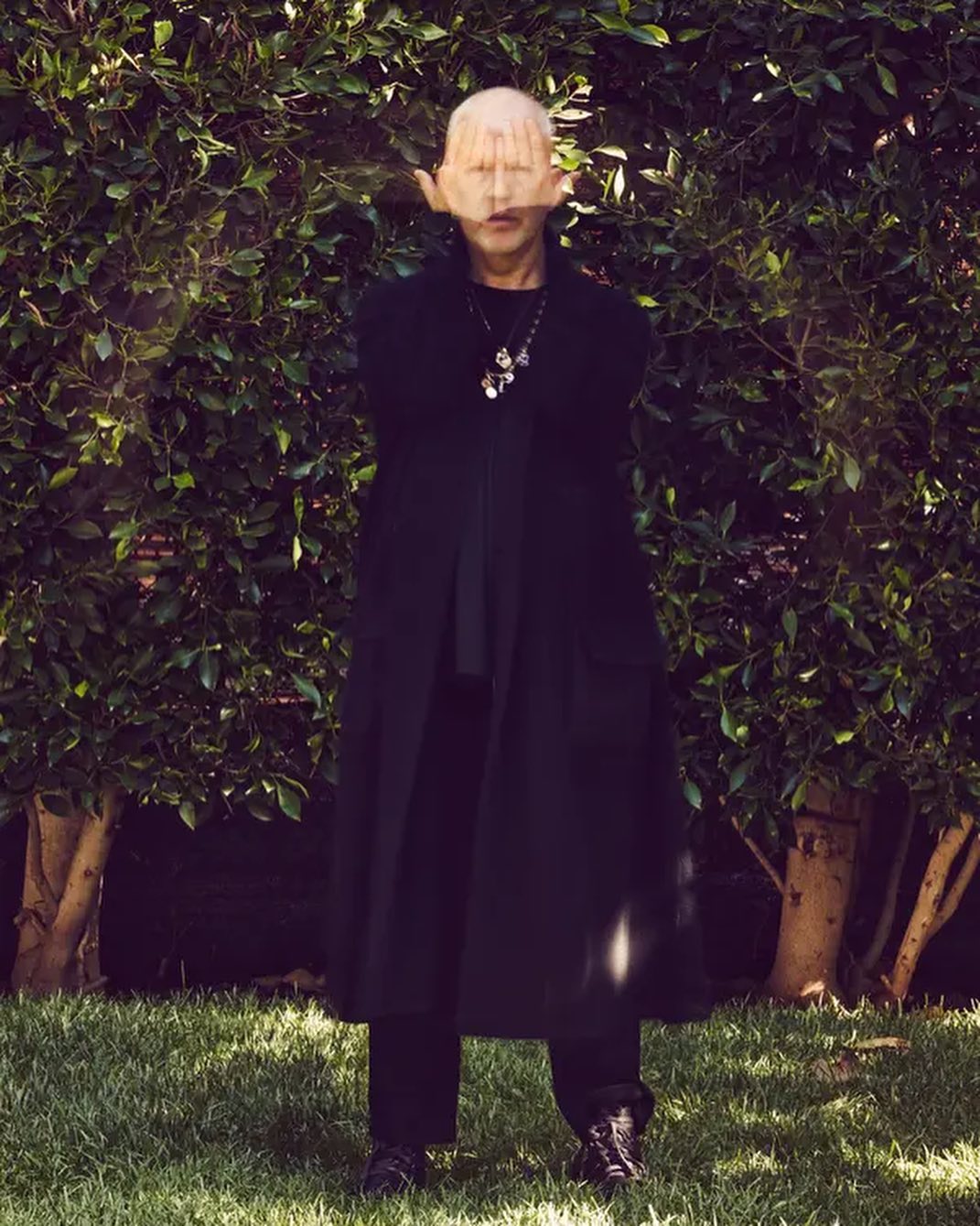 Mature adult in a long black coat stands in a backyard with palms covering their face, accessorized with a pendant necklace.