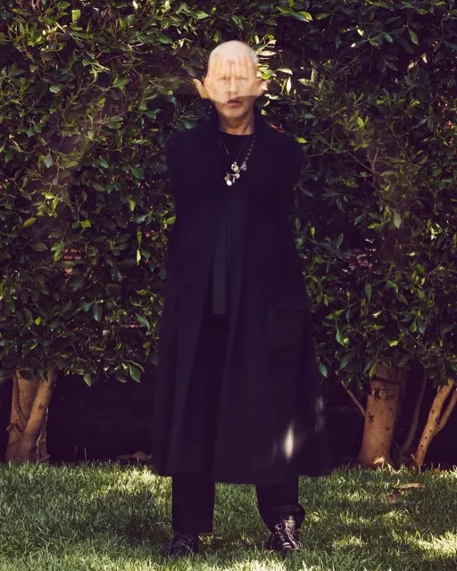 Mature adult in a long black coat stands in a backyard with palms covering their face, accessorized with a pendant necklace.