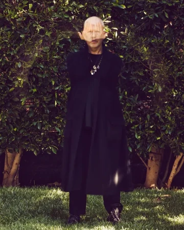 Mature adult in a long black coat stands in a backyard with palms covering their face, accessorized with a pendant necklace.