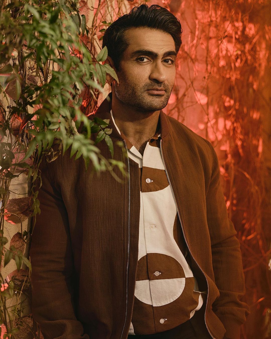 Portrait of Kumail Nanjiani, an adult male, wearing a brown jacket over a patterned shirt, posing in front of a green leafy backdrop.