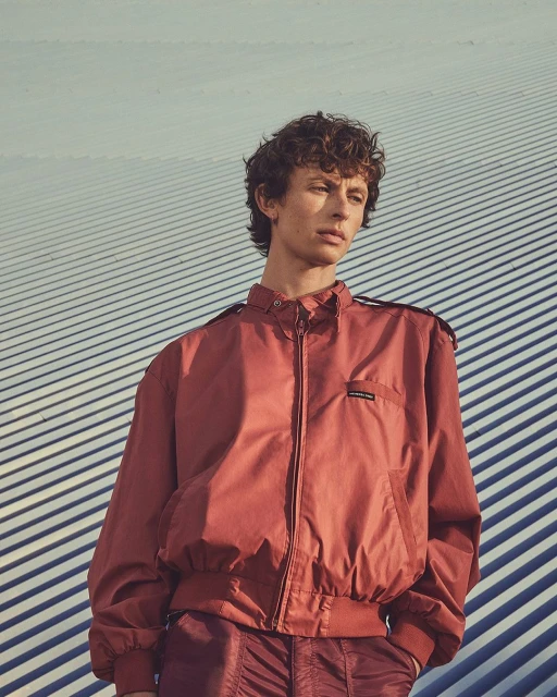 A young adult male stands outdoors wearing a reddish-brown jacket and pants against a striped background, gazing slightly off camera.