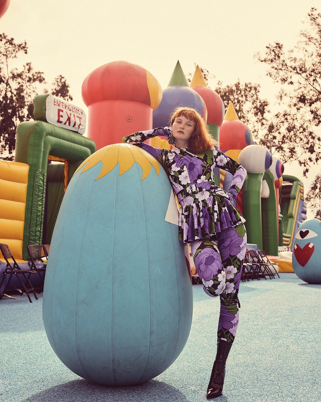 Adult woman in a floral pantsuit leans against a large inflatable egg in an outdoor setting with inflatable structures and chairs.