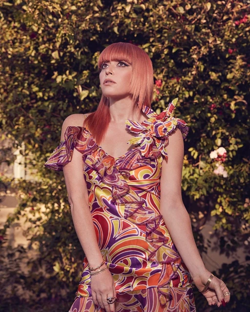 A woman with red hair wearing a retro patterned dress stands in front of leafy green foliage looking up and to the left.