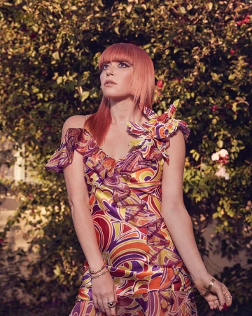 A woman with red hair wearing a retro patterned dress stands in front of leafy green foliage looking up and to the left.