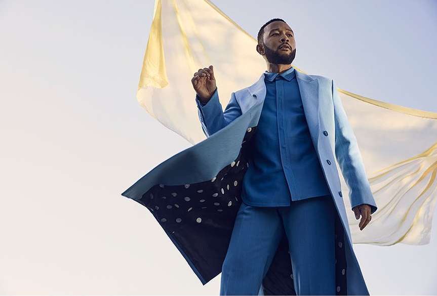 John Legend in a blue suit and coat poses against a light blue sky with a flowing yellow cloth backdrop, creating a fashion portrait.