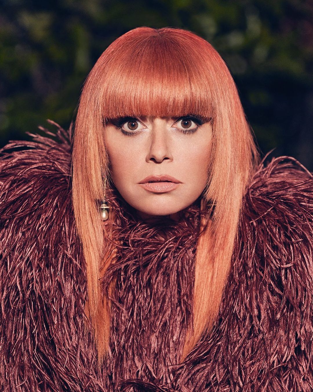 Portrait of a woman with copper-colored hair and bangs, wearing a burgundy feathered garment against a dark, blurred background.
