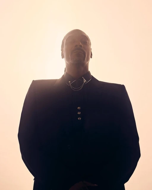 Portrait of Snoop Dogg in a dark suit against a muted orange background with low angle. The lighting creates a dramatic, silhouetted effect.