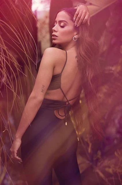 Elegant woman with long brown hair poses outdoors among lush greenery in a sleeveless black jumpsuit with an open back design.