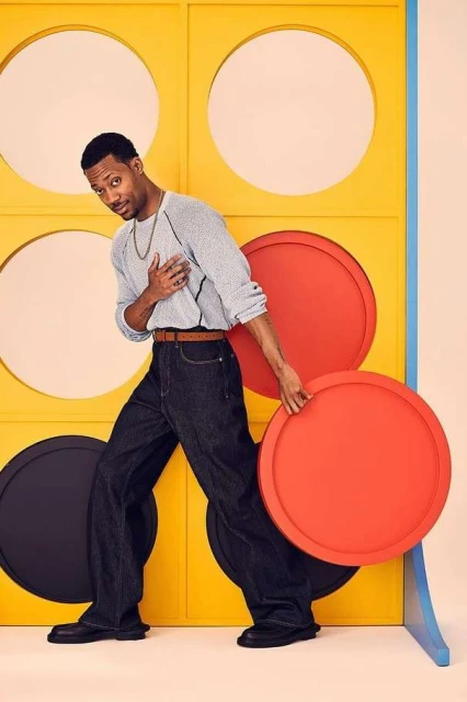 Stylish adult male in a grey sweater and dark jeans poses next to a yellow and red geometric prop in a studio with soft lighting.