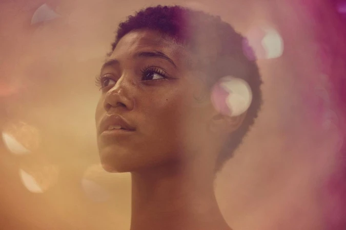 Close-up studio portrait of a young adult woman with short hair looking up; soft golden and pink light create a dreamy effect.