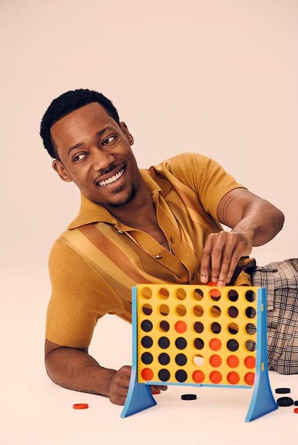 African-American man smiles while playing a Connect Four game on a white surface, wearing a striped short-sleeved shirt and checkered pants.