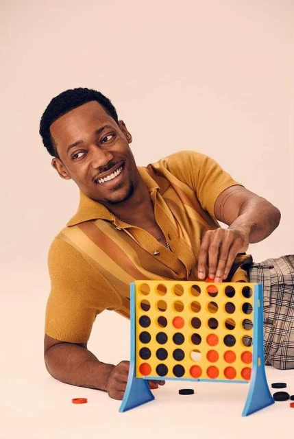 African-American man smiles while playing a Connect Four game on a white surface, wearing a striped short-sleeved shirt and checkered pants.