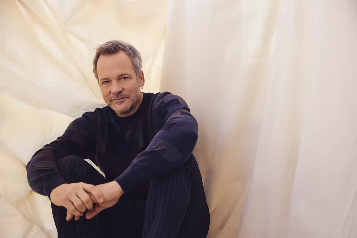 Mature man sits cross-legged with a thoughtful expression, hands clasped, wearing a dark blue sweater and pants against a cream backdrop.
