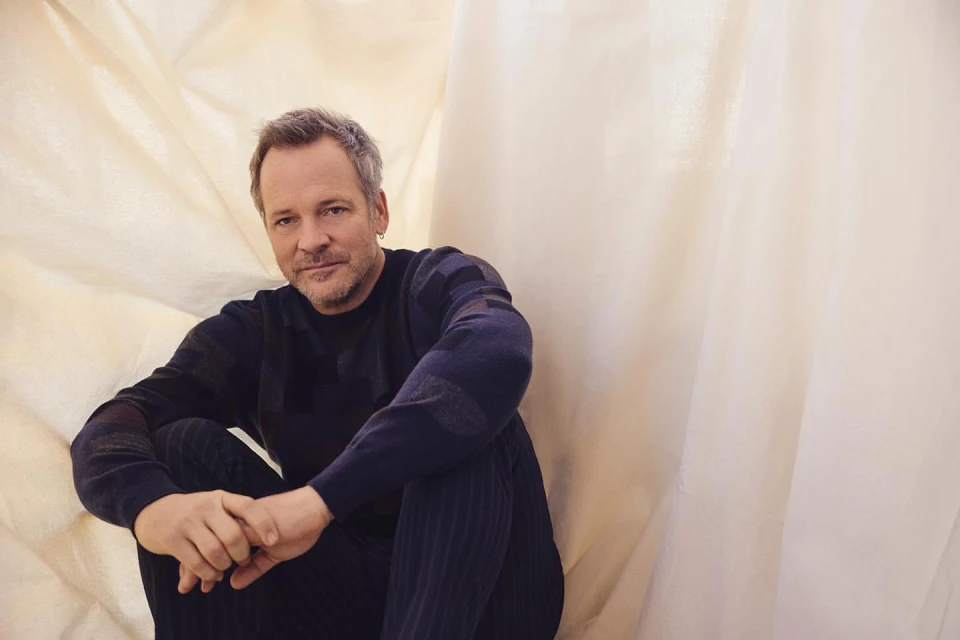 Mature man sits cross-legged with a thoughtful expression, hands clasped, wearing a dark blue sweater and pants against a cream backdrop.