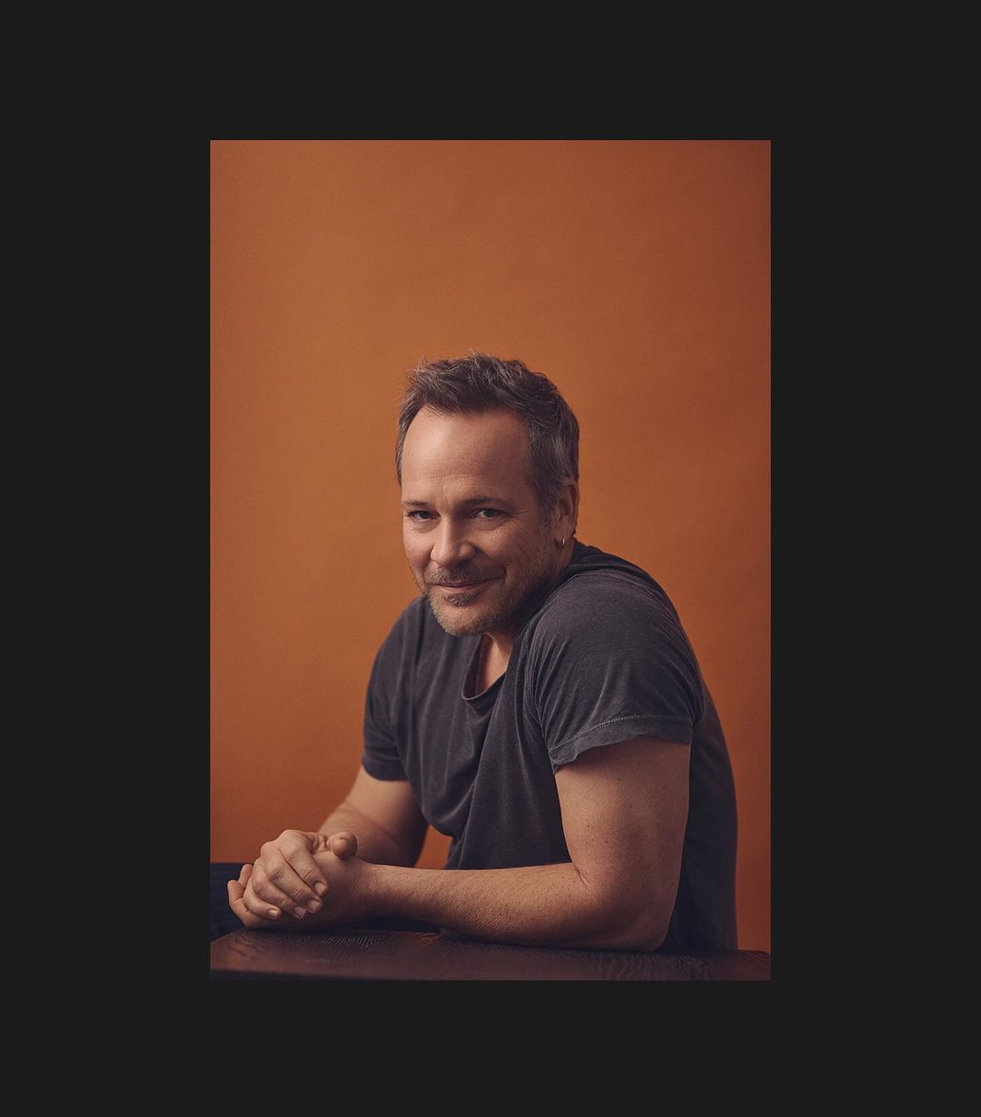Portrait of adult man with short hair and gray t-shirt in front of orange studio backdrop with hands clasped together on dark table.