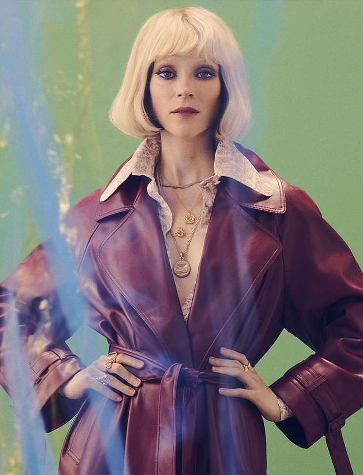 Blonde woman in a maroon leather trench coat stands against a mottled green background, illuminated by soft, diffused lighting.