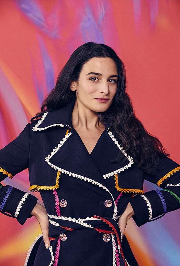 Adult female poses in a navy trench coat with decorative trim against a red and purple abstract backdrop in a medium portrait shot.