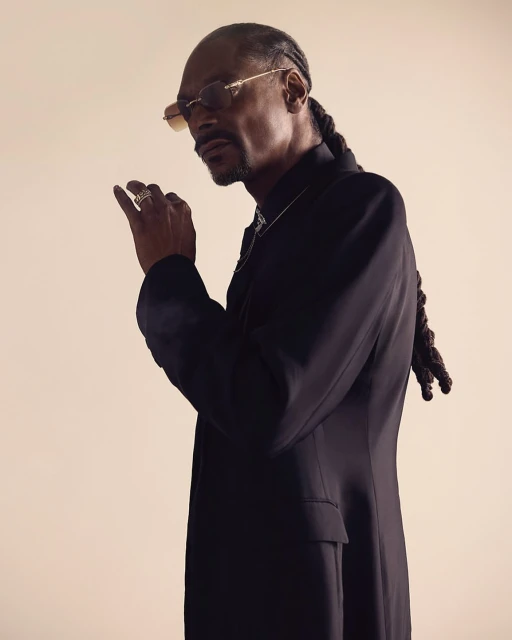 Portrait of Snoop Dogg in a black suit and sunglasses posing indoors against a cream background in a studio setting with rim lighting.