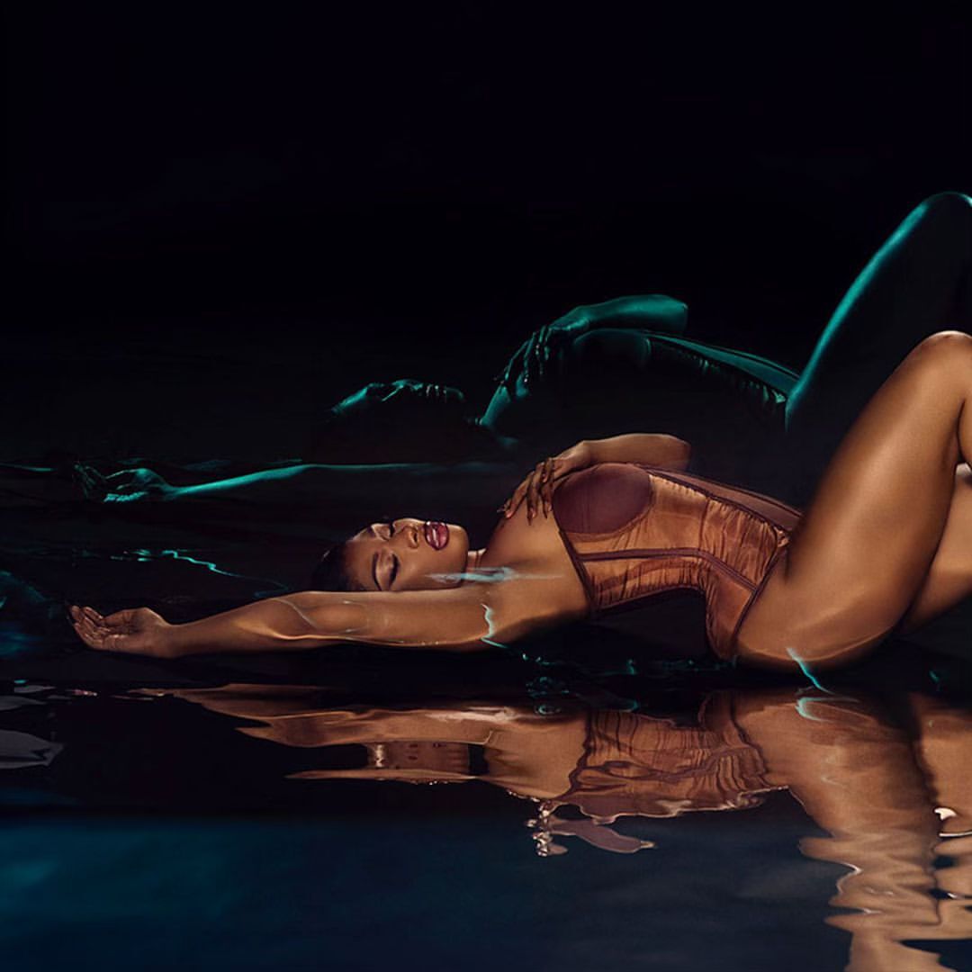 Woman in bronze corset and makeup lying in water with reflection against a dark teal background in a studio shot.