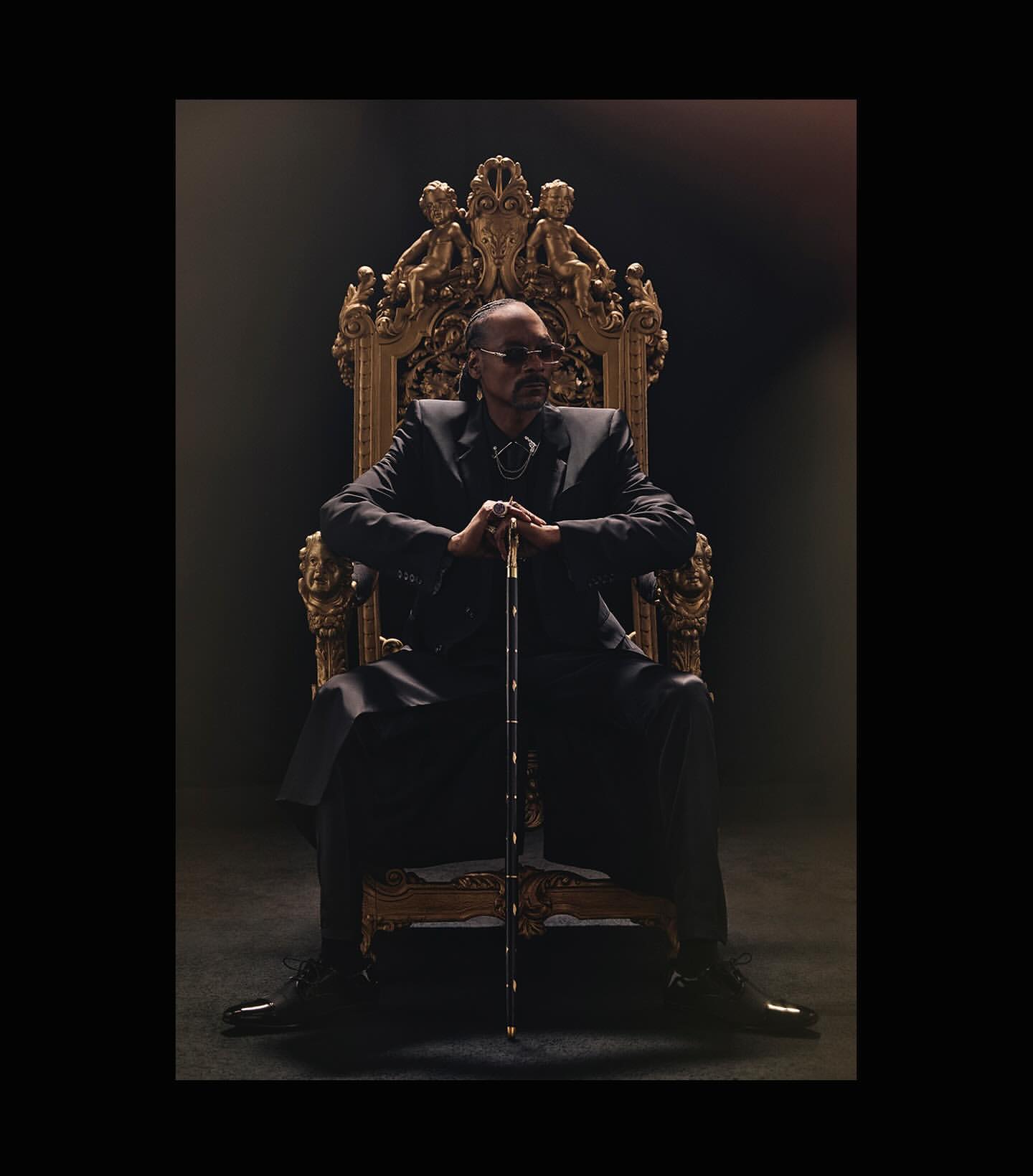 Snoop Dogg seated on an ornate gold throne, holding a cane, wearing a dark suit against a dark background in a regal portrait.