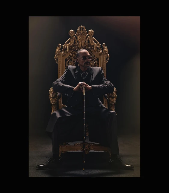 Snoop Dogg seated on an ornate gold throne, holding a cane, wearing a dark suit against a dark background in a regal portrait.